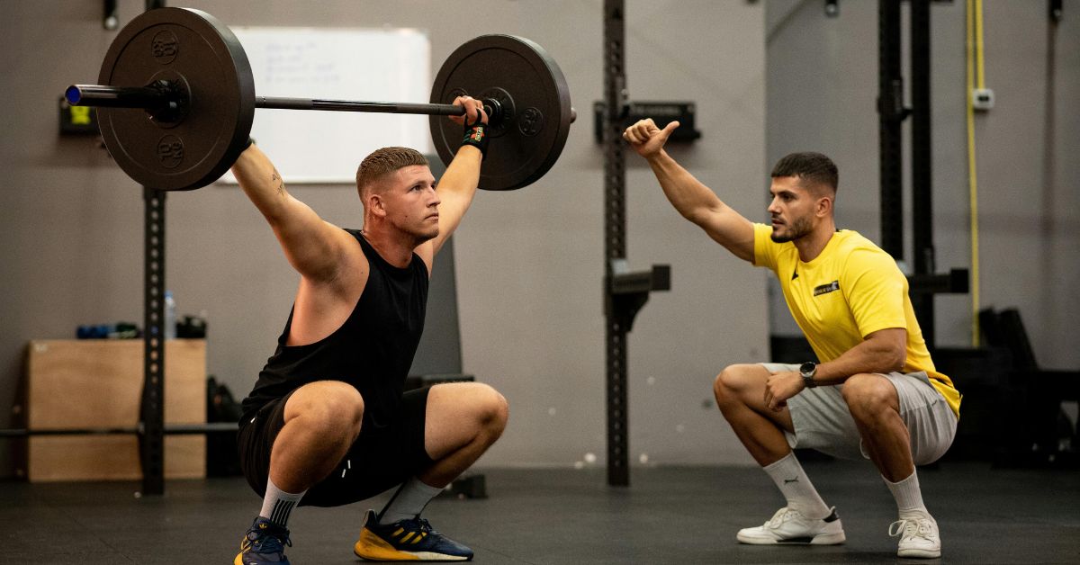A muscular man performing a weightlifting squat with a coach guiding him indoors.