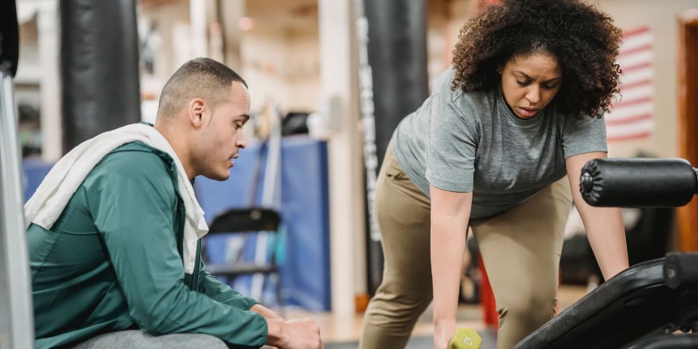 A personal trainer guides a woman lifting dumbbells in a gym setting.